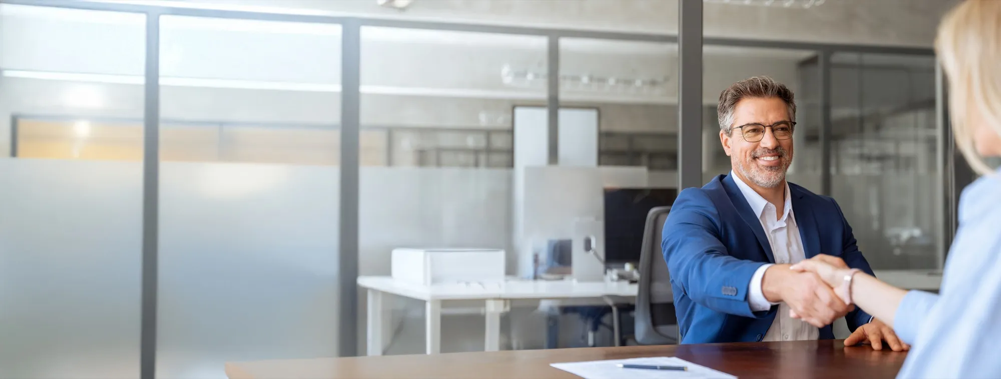 An attorney in a suit shaking hands with a client across an office desk.