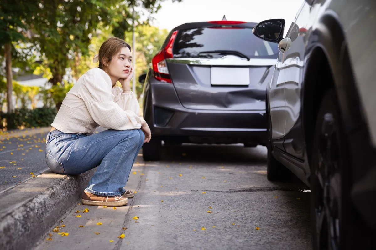 A young woman sits on a curb beside two cars after a collision.