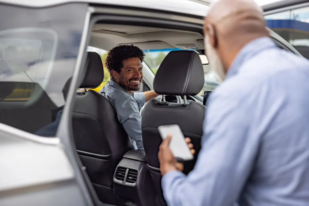 A rideshare passenger holding a phone speaks with the driver through an open car door.
