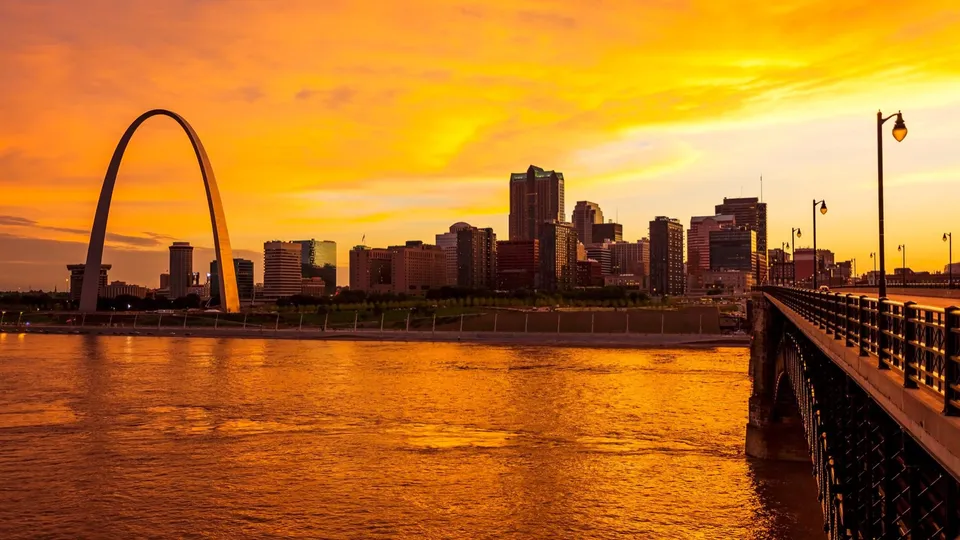 The Gateway Arch and downtown St. Louis skyline at sunset, seen across the Mississippi River from the Eads Bridge.